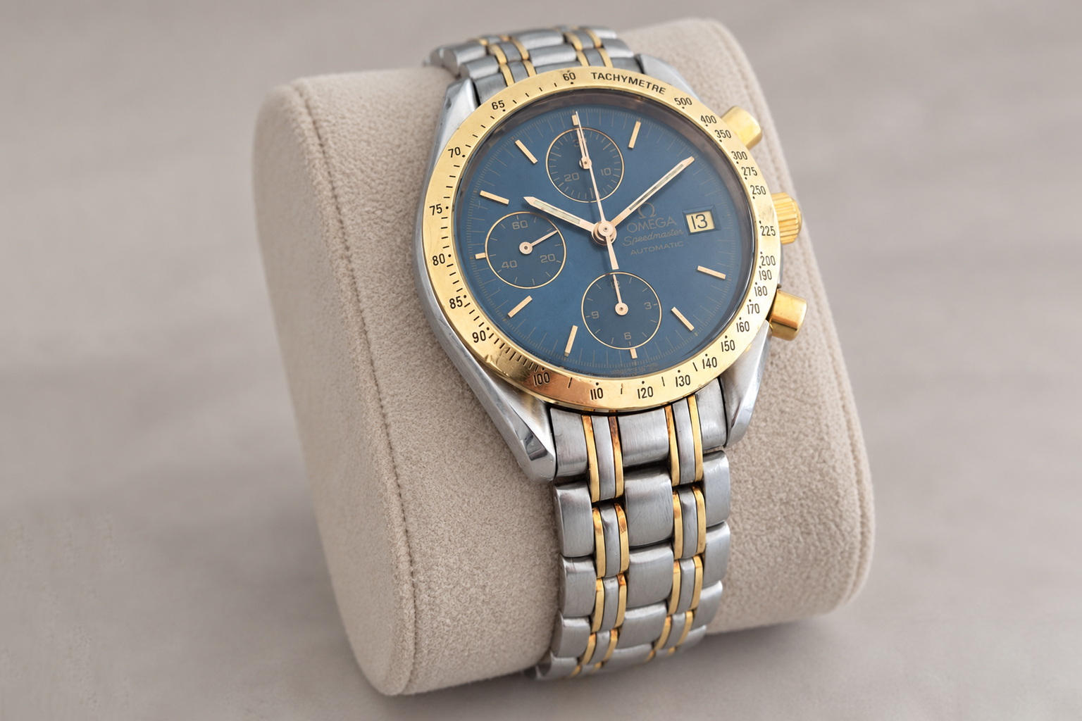 Reloj Omega Speedmaster de acero y oro, con esfera azul y brazalete bicolor, sobre un soporte de terciopelo beige.